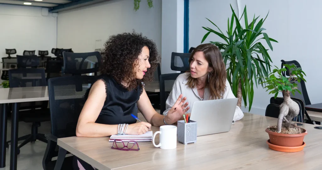 Two people in conversation at a desk with a laptop and notes