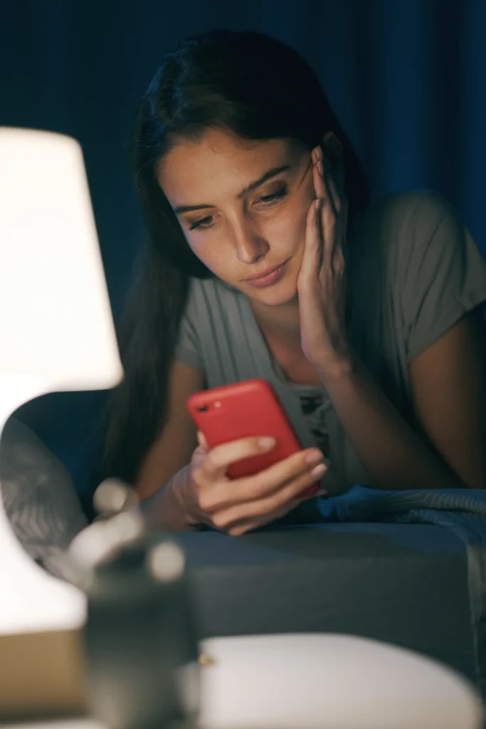 Woman lying in bed at night reading her phone by lamplight