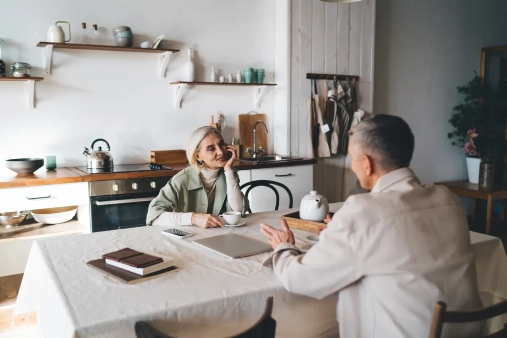 A woman with a laptop and notebook talking with a man seated across a kitchen table