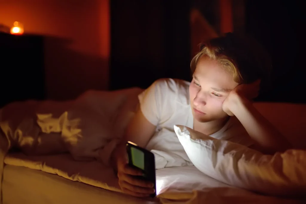 A teenager lying in bed at night with the glow of a phone screen on their face